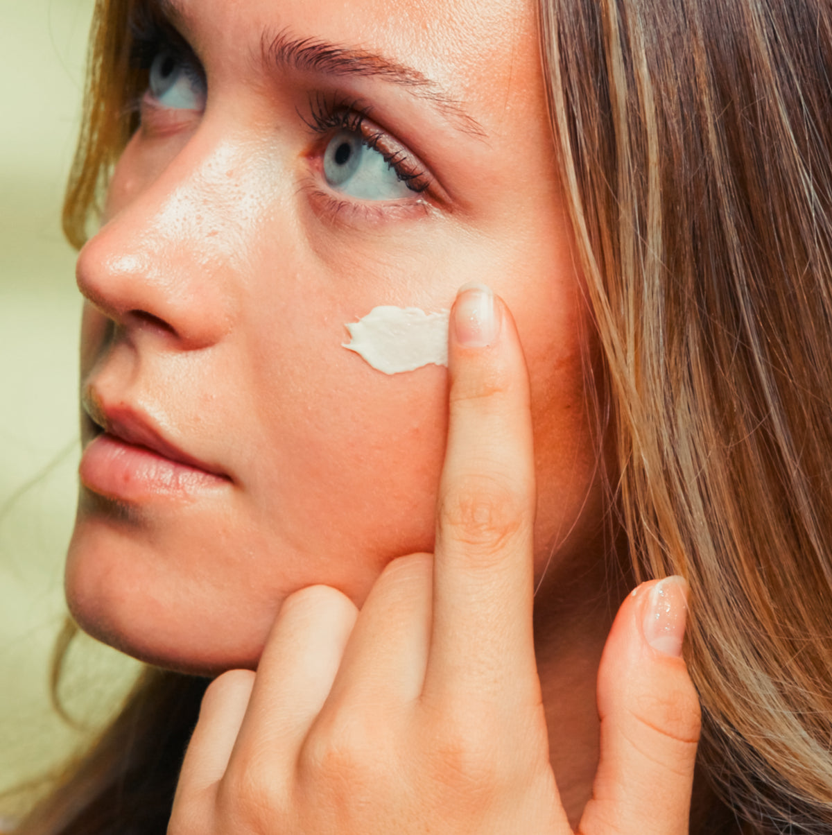 A woman pressing cream onto her cheek with one fingertip.