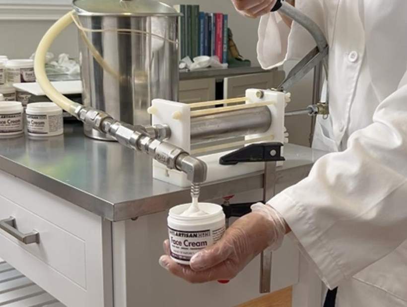 A gloved hand holding a jar under a small filling machine as cream is dispensed into it.