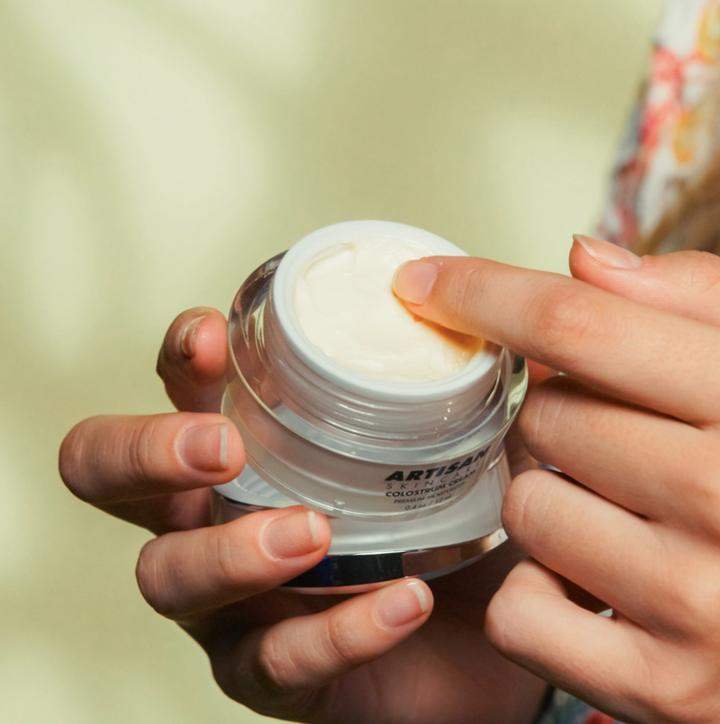 Hands holding the open Colostrum Cream jar, a fingertip lifting cream from the surface.