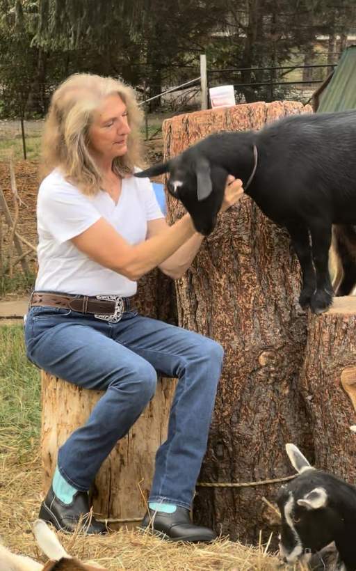 Lisa sitting on a log on the farm, one hand on a black goat that has climbed up beside her.