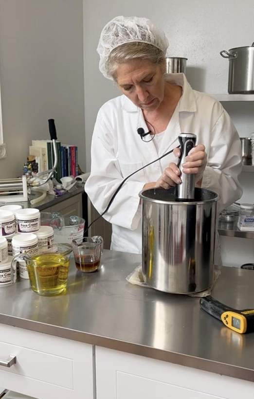 Lisa in a white coat and hair net, blending a batch of cream in a steel pot, with measuring cups of oils and rows of filled jars on the bench.