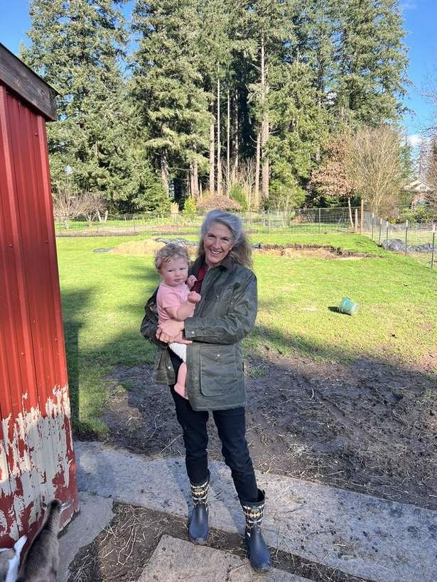 Lisa, the founder of Artisan The Goat, standing outside the red barn on the family farm in Southwest Washington, holding a small child, smiling.