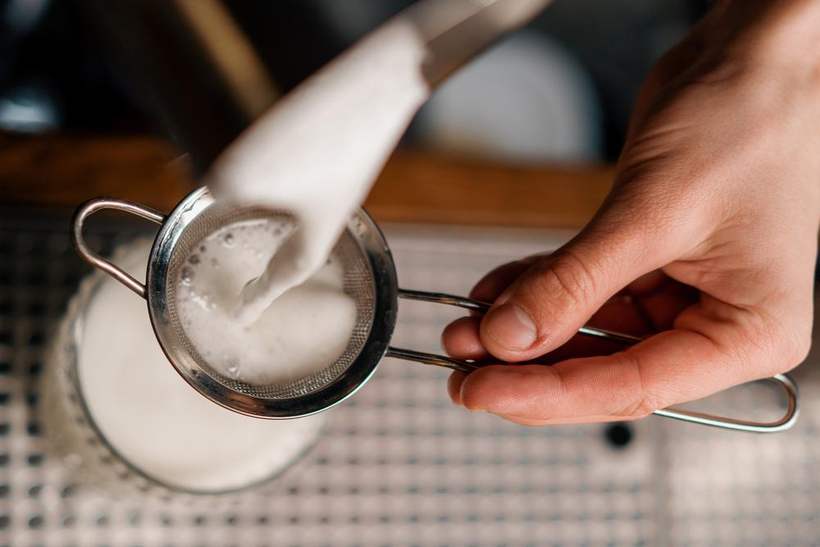 Fresh goat milk being poured through a fine strainer into a glass jar.